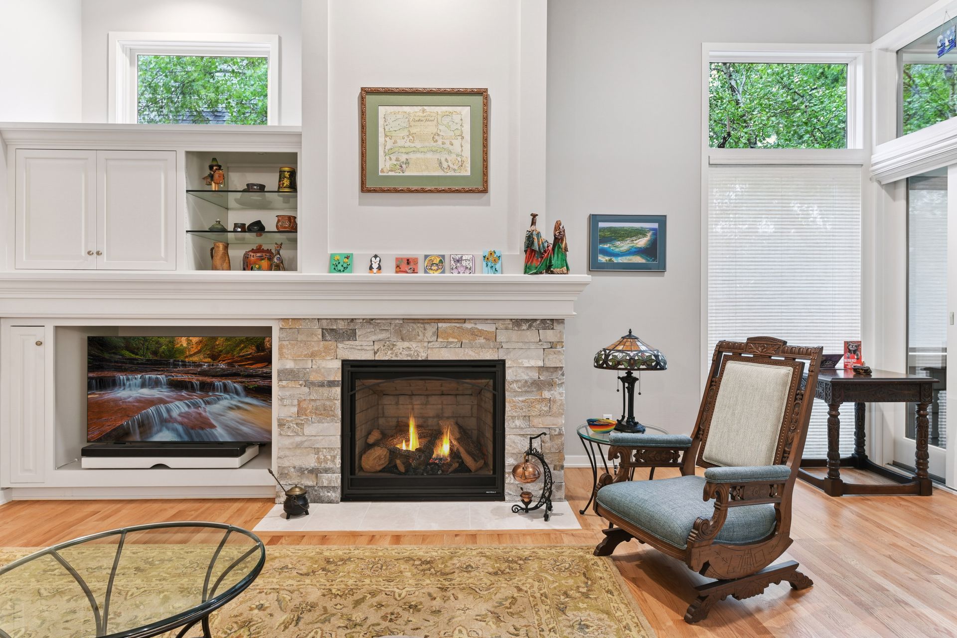 Traditional living room with a stone fireplace, built-in shelving, wood rocking chair, Tiffany-style lamp, and large windows providing natural light.