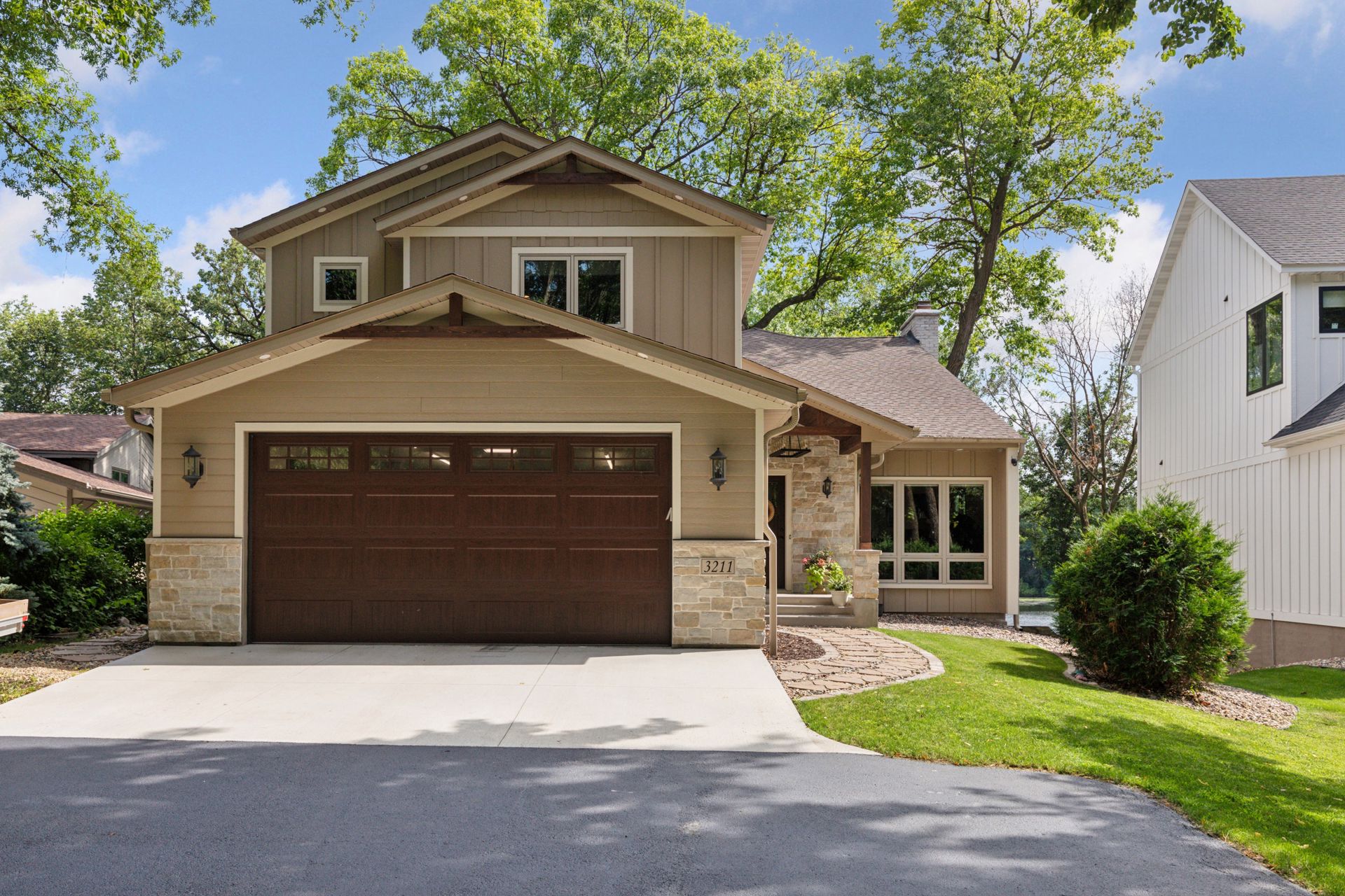 Two-story tan house with stone accents, dark wood garage door, and a covered front porch off a curved walkway.