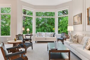 Bright traditional living room with large bay windows, white sofas, floral pillows, and carved wood armchairs facing a dark wood coffee table.