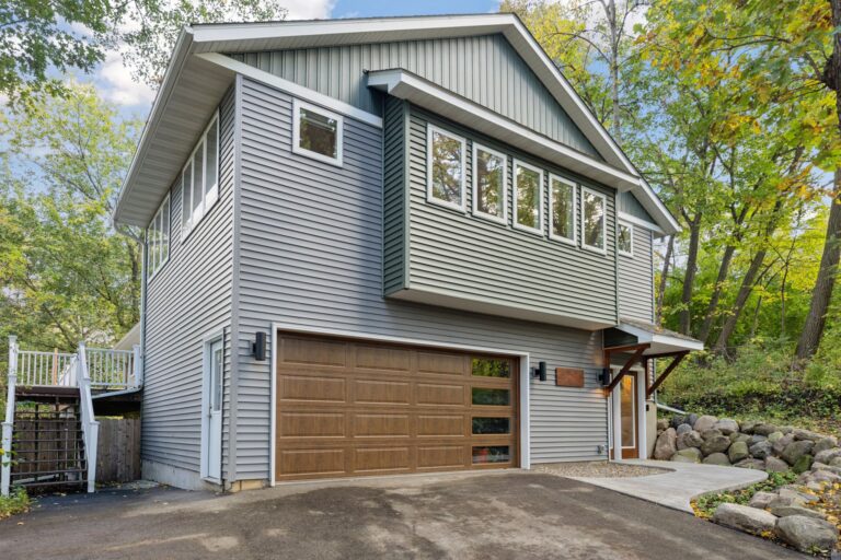 Modern two-story house with gray vinyl siding, a wooden garage door, and multiple rectangular windows on the upper level, surrounded by trees and greenery on a sloped driveway.